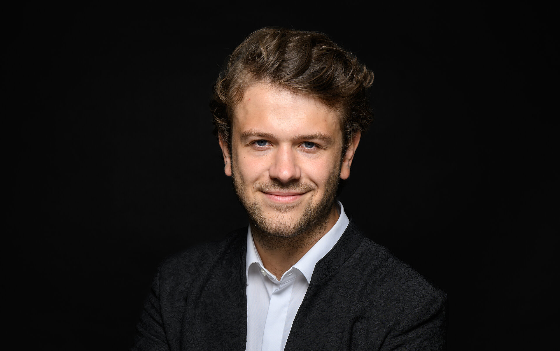 Portrait von Roman Lederer mit verschränkten Armen, dunklem Sakko und weißem Hemd, leicht lächelnd vor schwarzem Hintergrund.